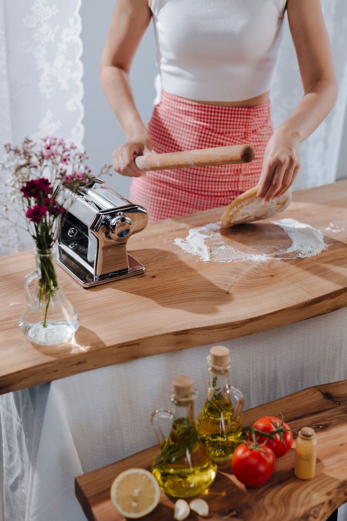 gallery-6 A woman preparing Italian pasta using a rolling pin and fresh ingredients in a cozy kitchen setting.