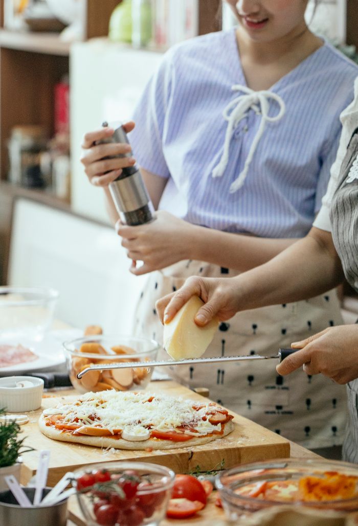 gallery-1 Crop anonymous females grating cheese on pizza placed on wooden board while cooking together in kitchen