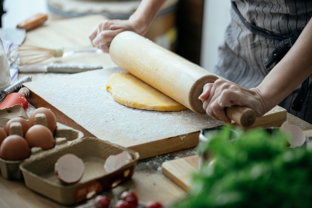 services-02 Unrecognizable female cook flattening dough with rolling pin while standing at table with wooden board against blurred background in kitchen