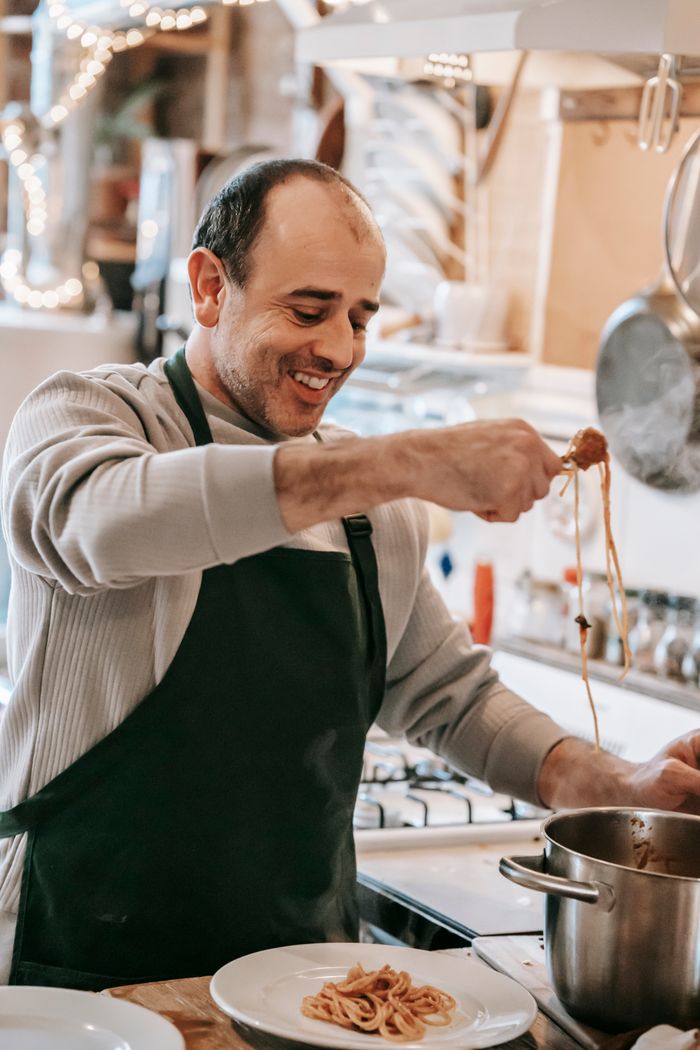 gallery-4 Cheerful adult ethnic male in casual clothes and apron smiling while putting appetizing saucy pasta from saucepan into plate standing at table in kitchen